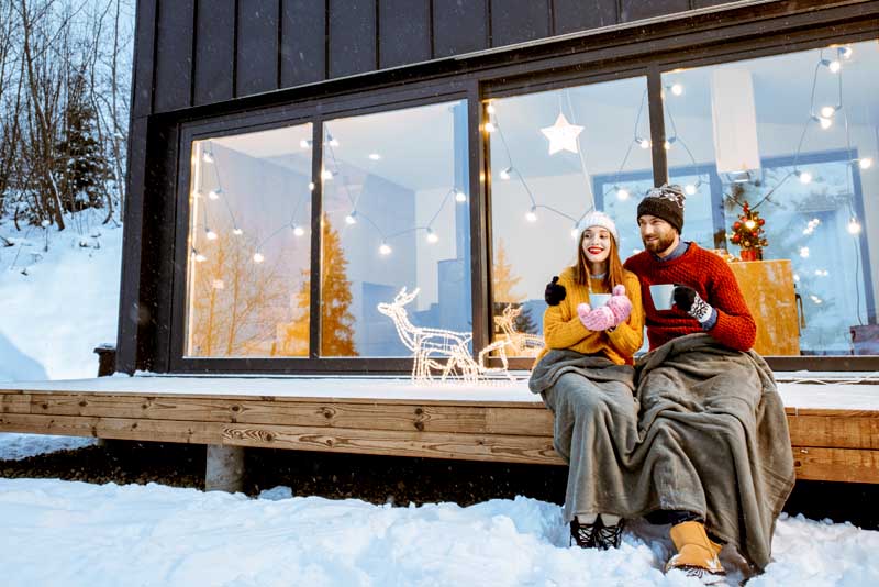 Image of a couple sitting outside a ski lodge under a blanket, drinking a hot drink.